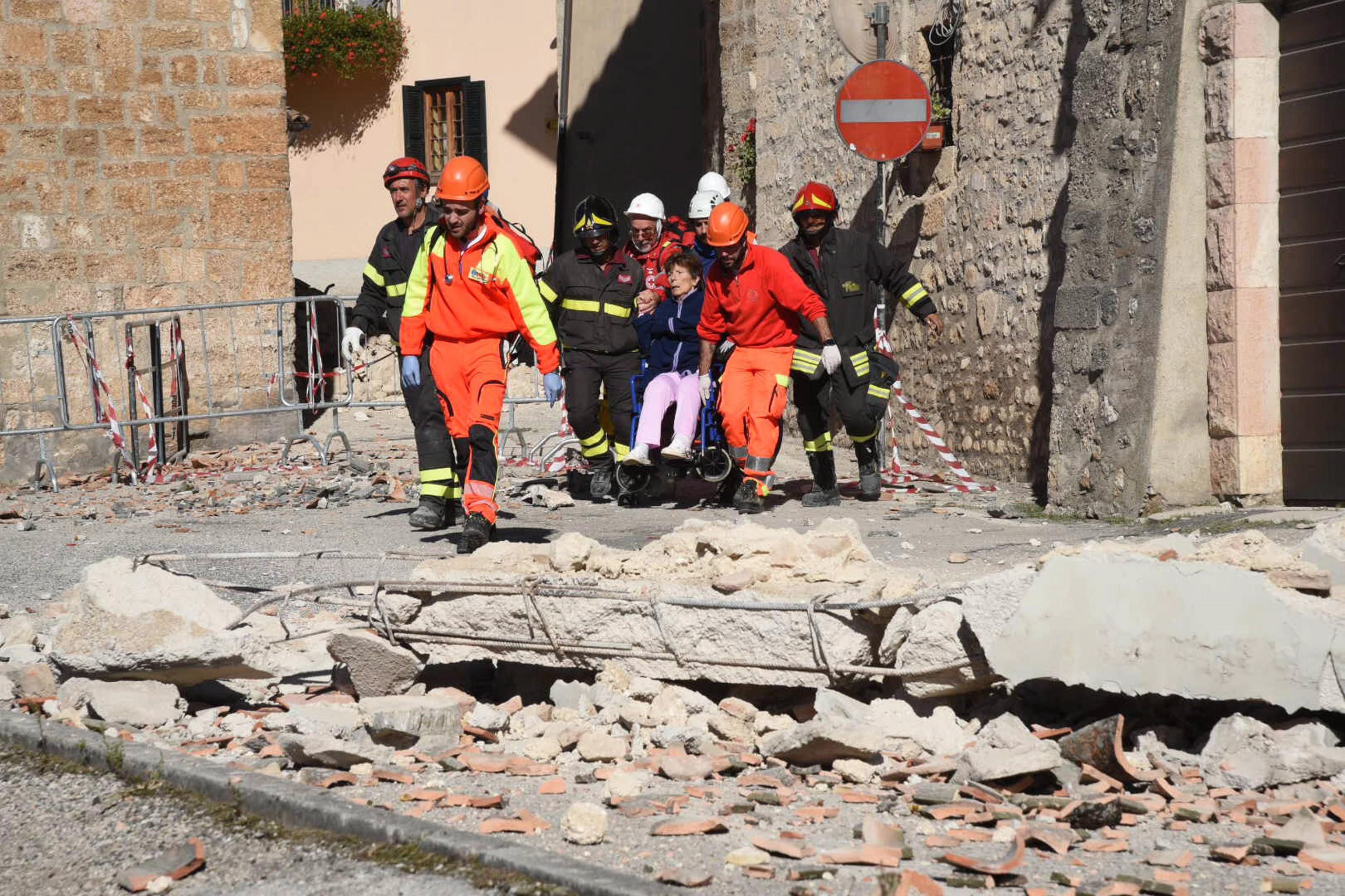 Norcia, 2016. október 30. Tûzoltók kísérnek egy kerekesszékes idõs nõt az utcán a közép-olaszországi Norciában 2016. október 30-án, miután a reggeli órákban Richter skála szerinti 6,5-ös erõsségû földrengés rázta meg a térséget. A földmozgás miatt, amelynek epicentruma Norcia és a tõle 20 kilométerre, északra fekvõ Preci között 10 kilométeres mélységben volt, súlyos károk keletkeztek és többtucatnyian megsérültek. Október 26-án három nagy erejû földrengés pusztított Olaszország középsõ régiójában. (MTI/EPA/Matteo Crocchioni)