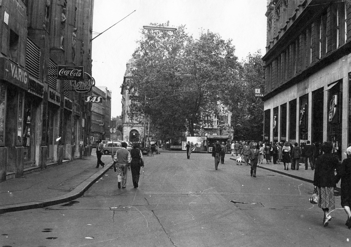 1980, a Váci utcáról sétálunk a Vörösmarty tér felé. Véletlen egybeesés, de a jobb oldali sarkon most is óraszalon van, a balon viszont már a Váci1 áruház, amiben nemrég nyitott meg a H&M. Akkortájt még autóval is be lehetett hajtani a Vörösmarty térre, ami parkolóként működött. Fotó: Fortepan.hu